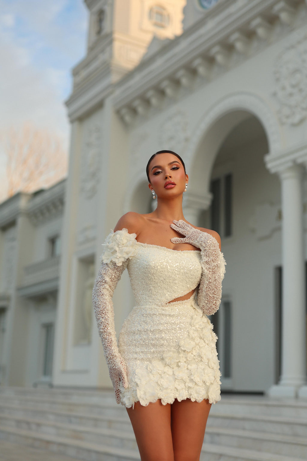 Woman in a white dress with floral details and long gloves standing in front of an elegant building.