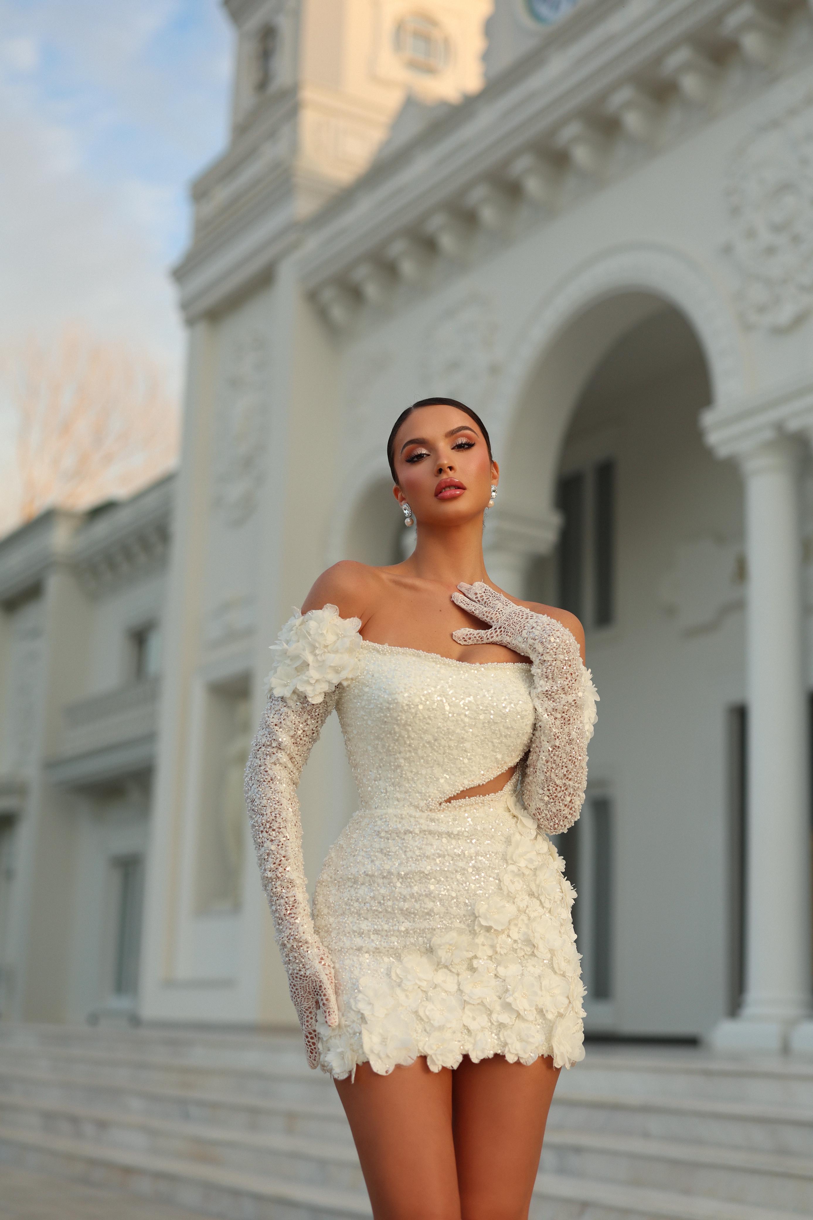 Woman in a white dress with floral details and long gloves standing in front of an elegant building.