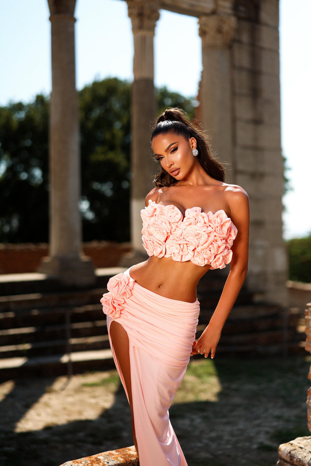Woman in a pink floral top and skirt standing in an outdoor setting with columns.