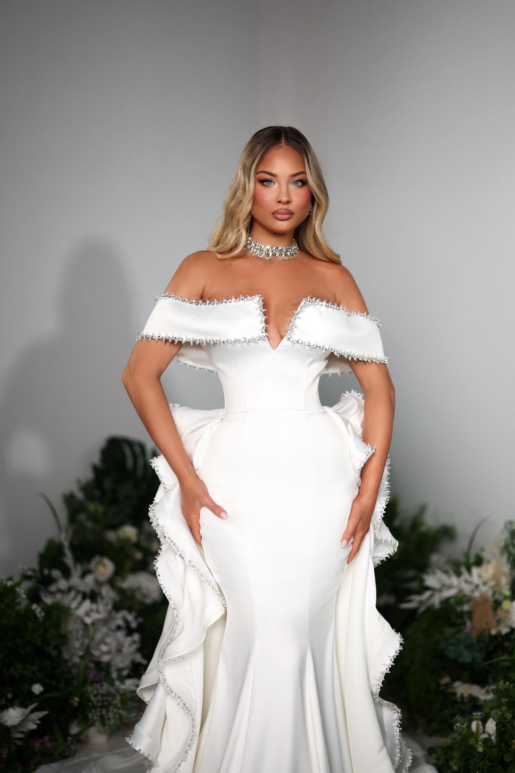 Woman wearing a white off-shoulder gown with ruffled details against a neutral background.