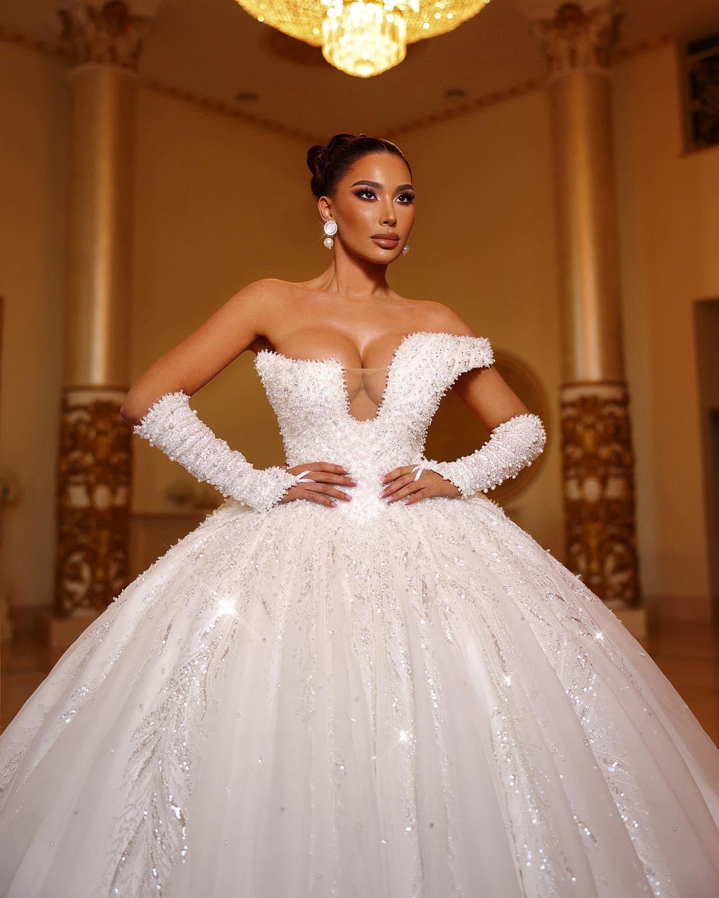 Woman in a white wedding dress standing in an elegant indoor setting with chandelier.