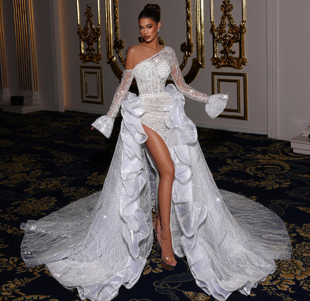 Woman in a white, one-shoulder gown with a high slit standing in an ornate room.