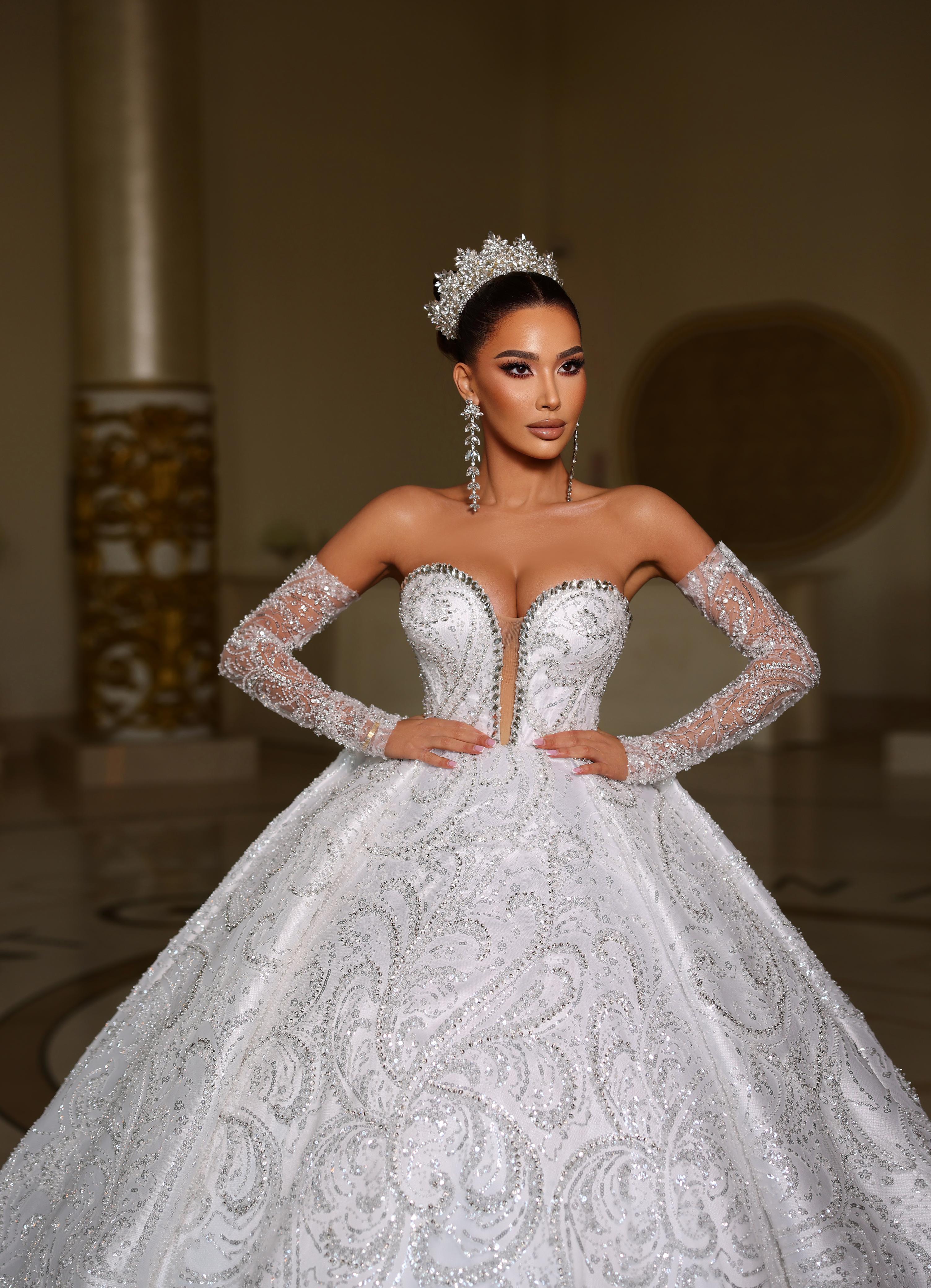Woman in a white wedding dress with intricate detailing and long sleeves, standing indoors.