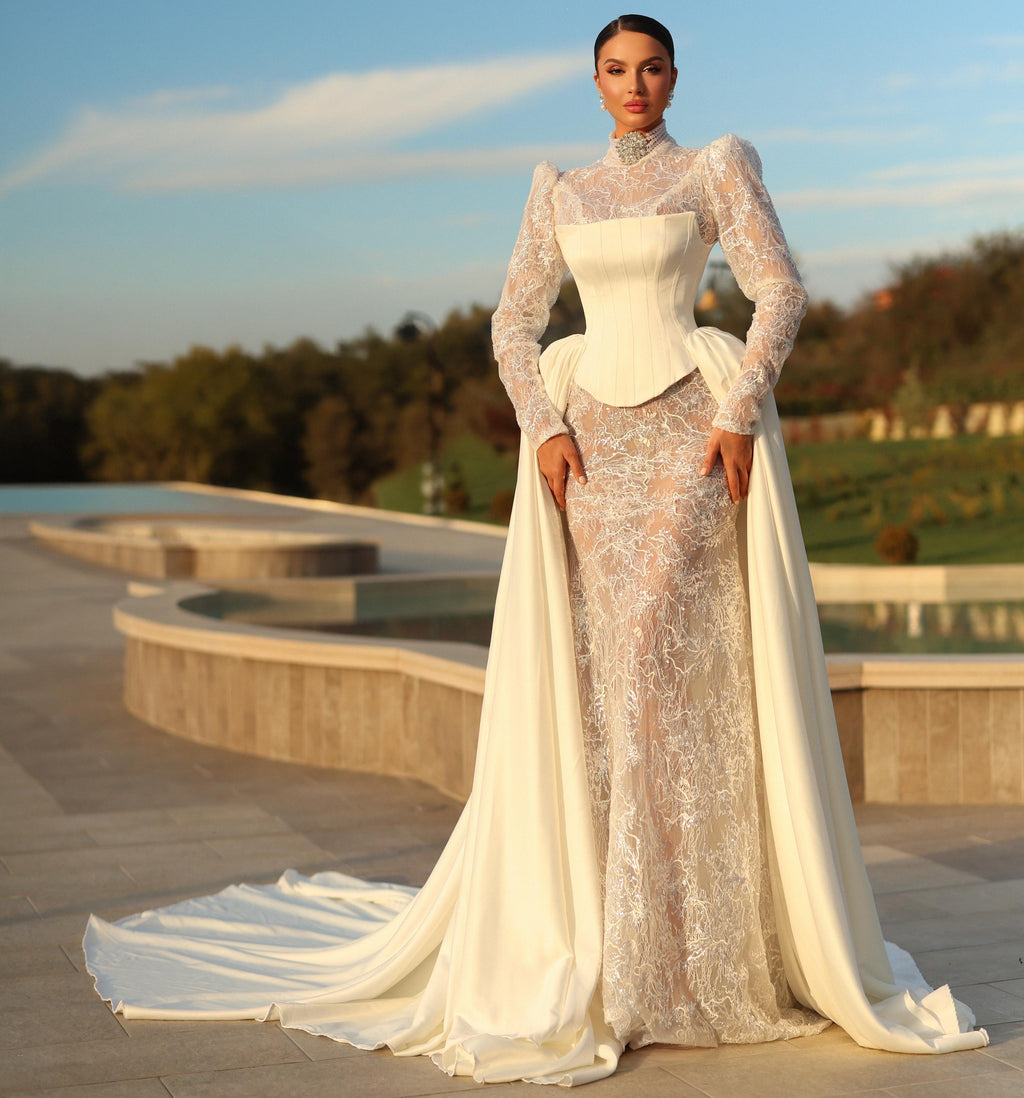Woman in a white lace gown standing outdoors with trees and sky in the background