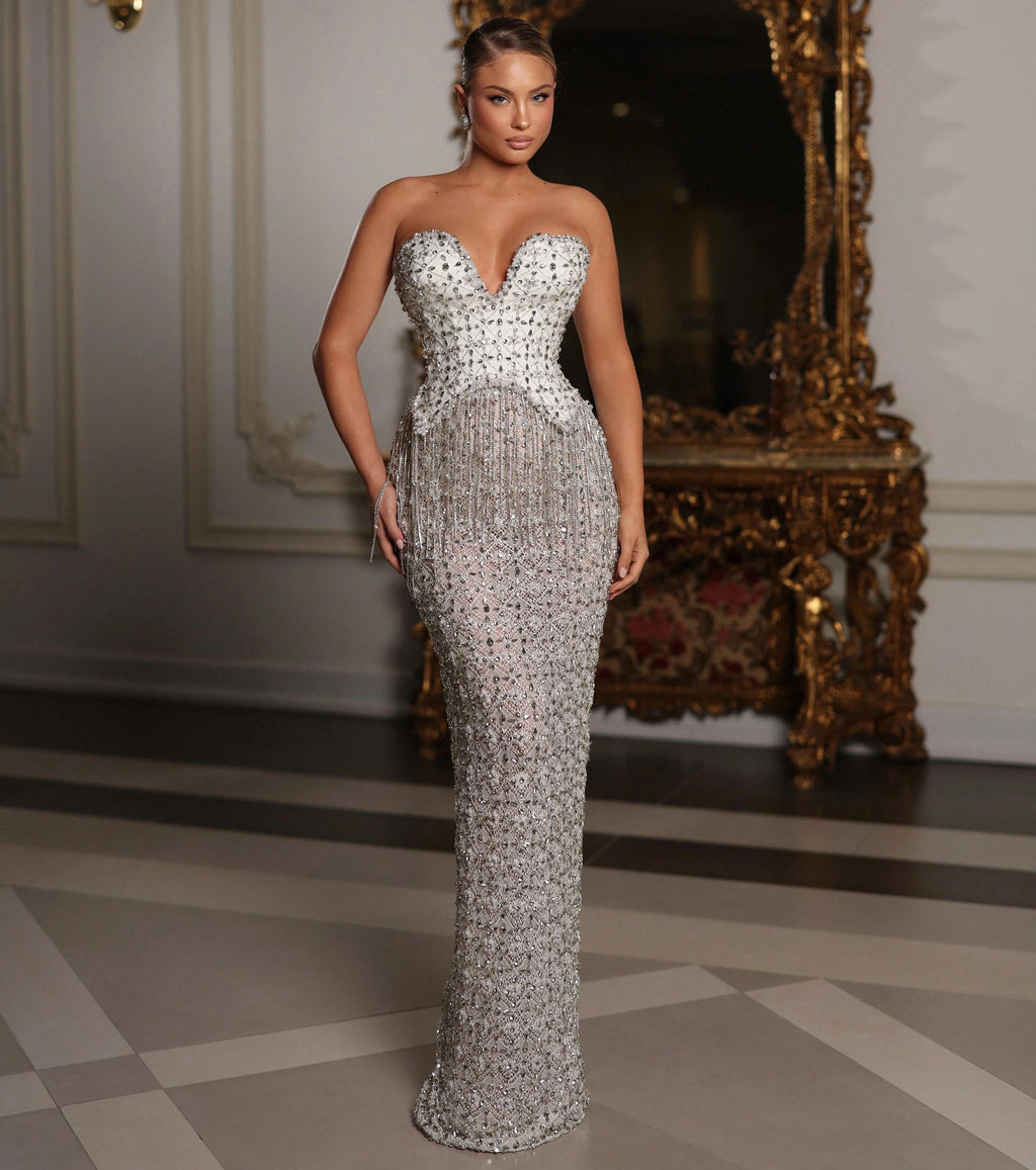 Woman in a silver strapless gown standing in an elegant room with a decorative mirror.