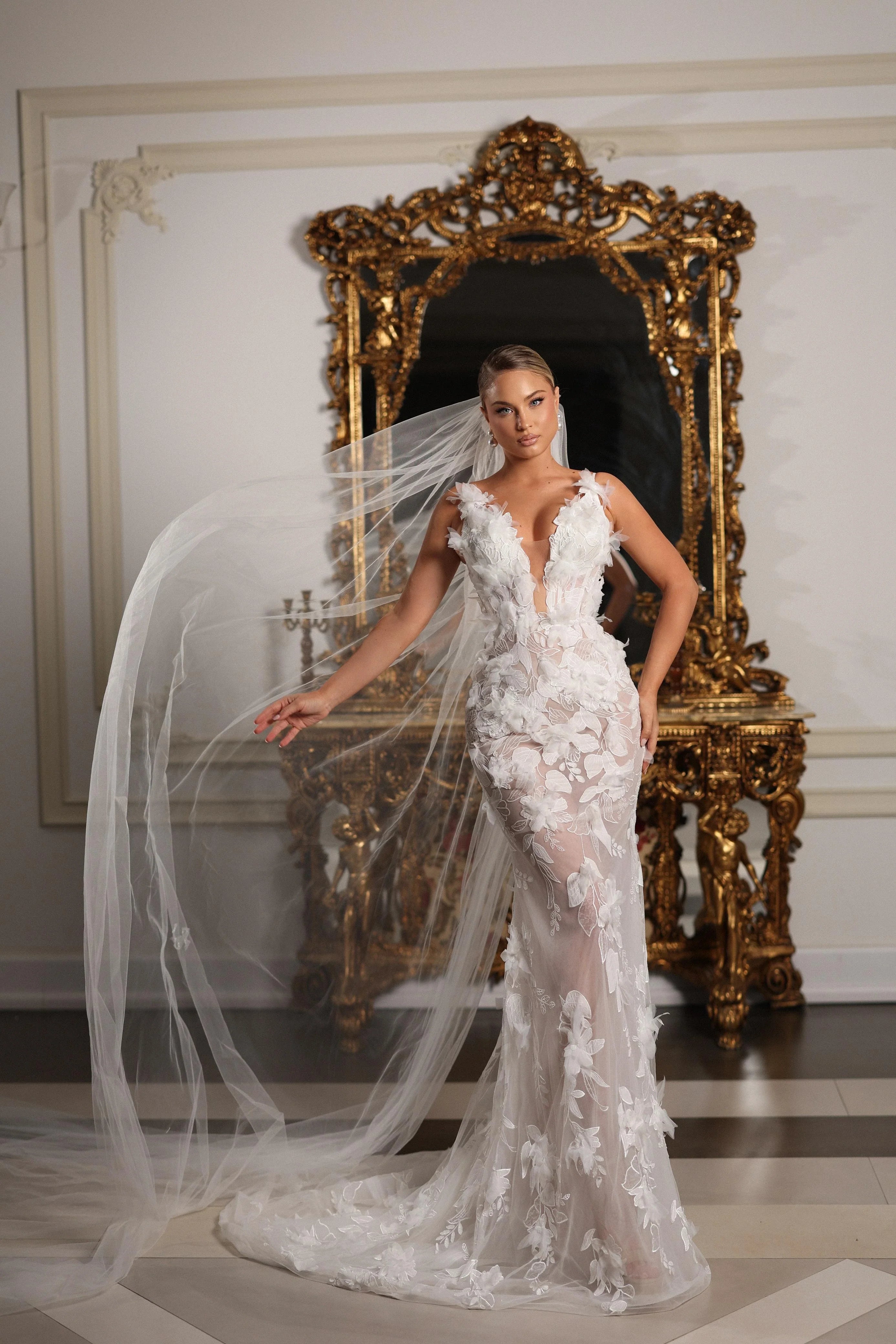 Woman in a white wedding dress with a long veil standing in front of an ornate mirror.