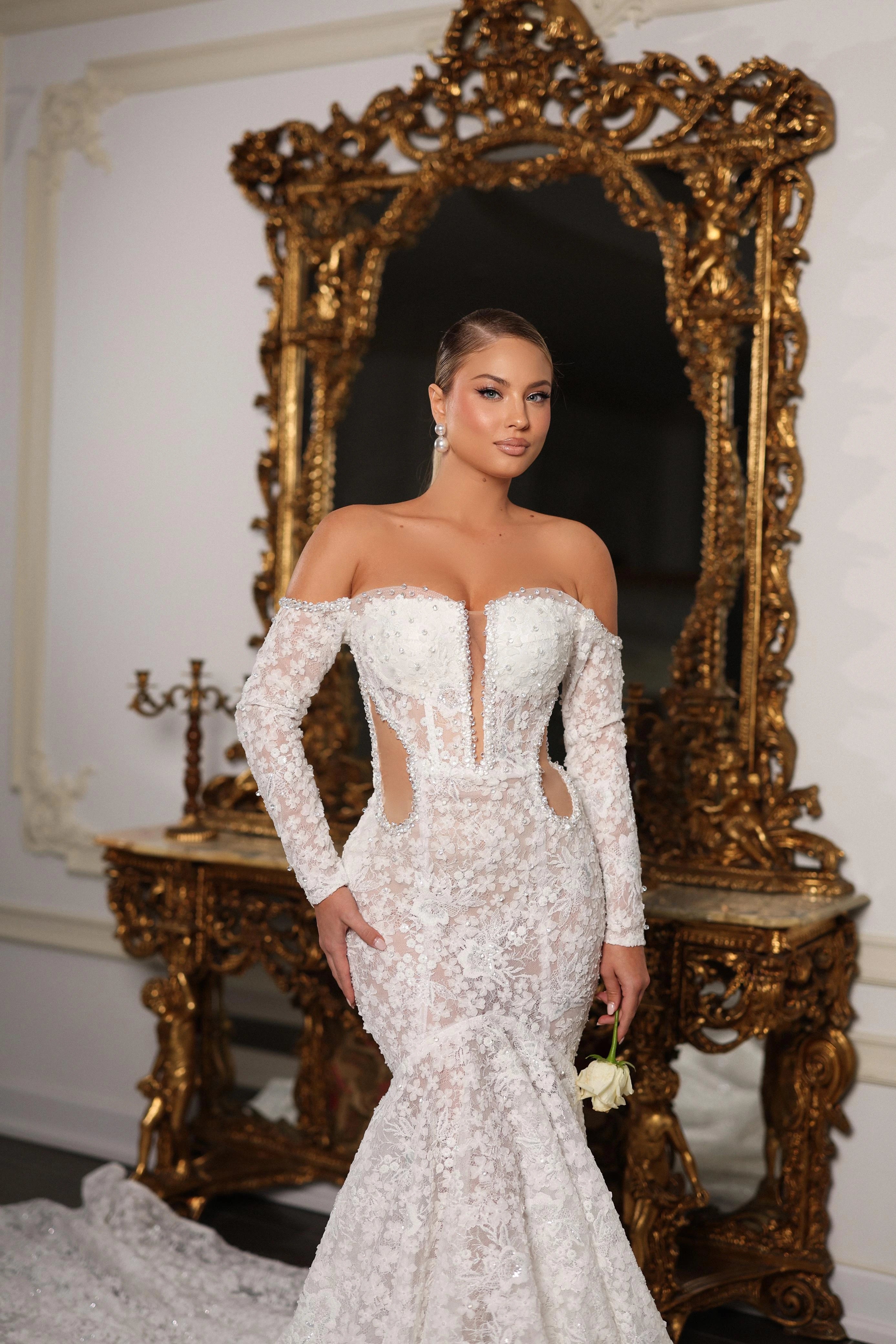 Woman in a white lace wedding dress standing in front of an ornate mirror.